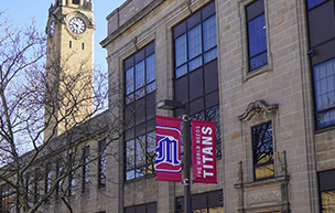 A photo of the Engineering Building near the clock tower with red banners that feature the °ϲ logo and World Needs Titans on it.